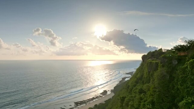 Tropical Bukit Peninsula coastline at sunset, green cliffs and rolling waves with a paraglider soaring over the horizon during golden hour adventure. Travel and explore Bali nature and activities