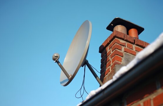 Satellite dish installed on brick chimney for internet access. Device receives signal for home broadband connection. Outdoor technology on roof with clear blue sky.