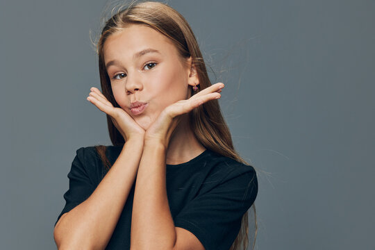 Girl with long hair making kiss face with hands near cheeks posing on gray background. Portrait of young female child showing playful and cute expression in studio.