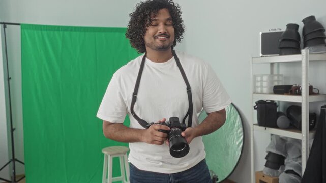 Man photographer holding dslr camera with strap, hands on lens, smiling in photo studio with green screen, stool and reflector; creative joy.