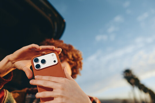 Woman with curly hair wearing rainbow sweater taking photo with smartphone outdoors near car, smiling and enjoying lifestyle moment under clear sky with palm trees in background