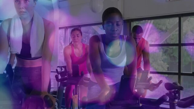 Pedaling center female in white tank top leaning on handlebars at gym, with bikes towel bokeh
