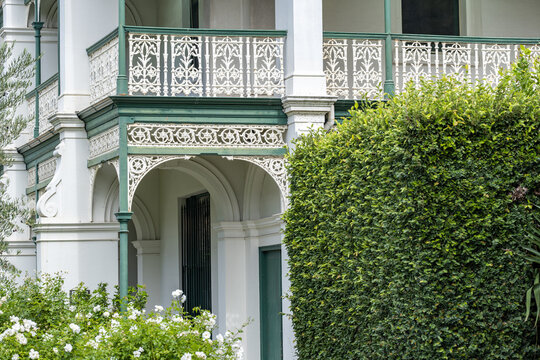 Ornate heritage veranda and balcony of a Victorian-era house in Melbourne, Australia, featuring intricate iron lacework and lush garden hedging. Upscale residential architecture, leafy private living.