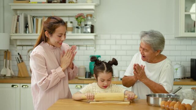 Happy Asian multi-generational family cooking together in kitchen. Little daughter rolls dough with rolling pin while mother and grandmother cheer and give thumbs up during baking session at home.