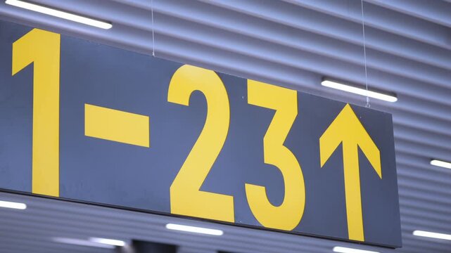 Airport gate sign shows numbers 1-23 with upward arrows. Sign is dark blue with bright yellow text. Ceiling has striped panels and fluorescent lights