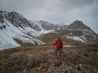 Fototapeta premium Male hiker in a red jacket with trekking poles standing on a rocky ridge against a backdrop of snowy mountain peaks under a cloudy sky, adventure travel and hiking concept.