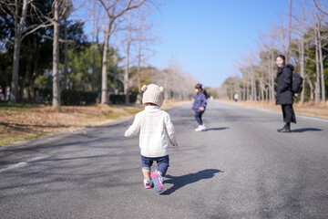 Fototapeta premium Sunlight glows warmly over a quiet park path. Child in a knit hat runs joyfully toward the distance. Another child plays ahead, barefoot and carefree. An adult watches from the side, smiling gently