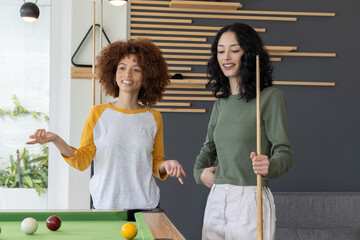 Obraz premium Two female friends playing pool in raglan and olive tops, smiling and holding cue with balls
