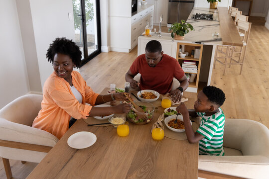 African American family with child sitting at wood table in modern kitchen, sharing pasta bowls