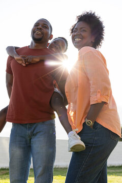 African American family standing on grassy shore, father carrying son on back, seawall, lens flare