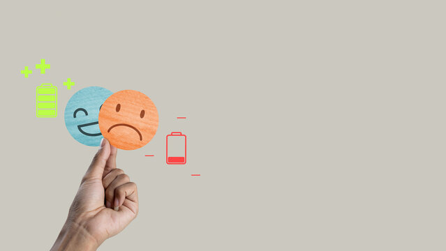 Conceptual image of emotional health featuring a hand holding a sad emoji over a happy one. Icons of full and empty batteries illustrate energy levels and mental exhaustion