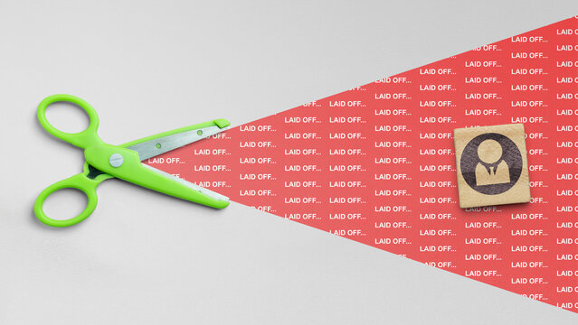 A conceptual top-down photo showing green scissors cutting into a red field with laid off text, alongside a wooden block featuring a human icon, representing job loss and downsizing