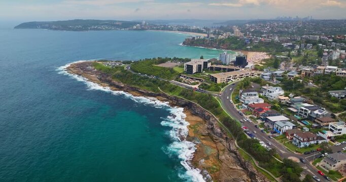 Sandstone coastline of Sydney's North Head, with turquoise ocean waves crashing against cliffs and the distant Manly beach, suburbs city skyline. Modern coastline cityscape aerial drone view flight