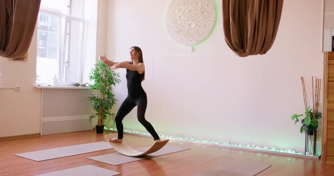 Fitness training. Woman workout. Athlete in black sportswear balancing on wobble board lifting arms for stability body skill in gym.