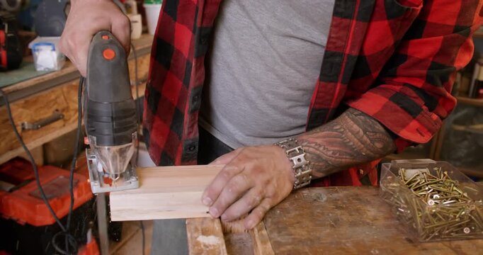 Joiner work. Furniture making. Male hand with professional jigsaw cutting wooden plank corner in curve line on workbench with sawdust.