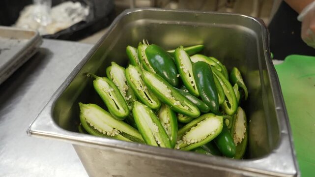 Prep cook slices jalapeno down the middle and tosses into stainless pan filled with halved peppers, slow motion close up 4K