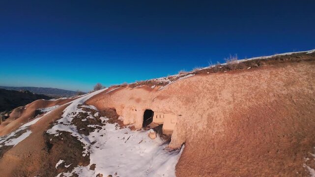 FPV drone flight past snowy cave entrances and carved tuff hillside in Uchisar, Cappadocia, Turkey, revealing winter terrain, ridge paths and a wide panoramic valley view. Pigeon valley, Turkiye