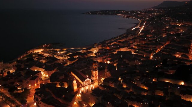 Menton old town at night, city lights of French Riviera stretching along dark Mediterranean Sea, France, drone