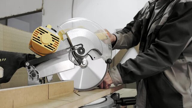 Worker using miter saw to cut wooden panel during woodworking process in industrial workshop with professional equipment.