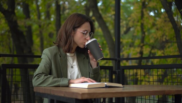 Young woman in a green blazer reads a book on a cafe veranda by the forest