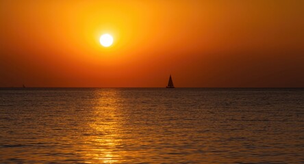 Fototapeta premium Golden sunset reflecting over calm ocean waters with distant sailboat silhouette