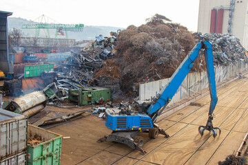Blue hydraulic crane lifting scrap metal at a recycling facility with large piles of metal debris and shipping containers in the background