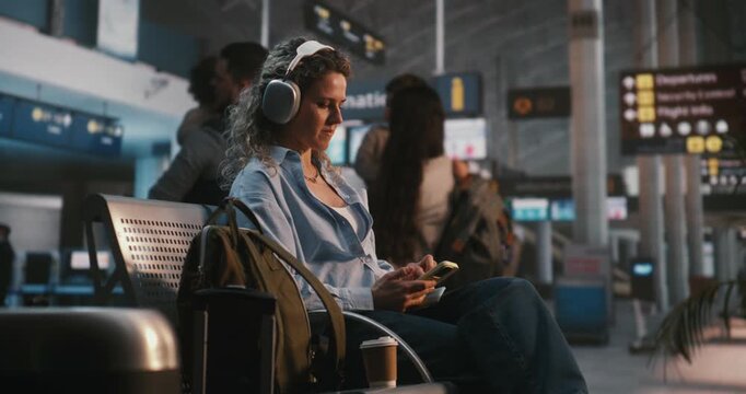 Stylish Female Traveler Enjoys Favorite Playlist and Browses Digital Content on Smartphone at Airport Lounge, Wearing Audio Headphones. Concept of Youth Travel Culture and Tech Savvy Lifestyle.