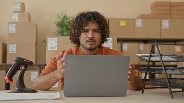 Man resting chin on hand while sorting papers and using laptop amid stacked cardboard boxes in a building; small business frustration.