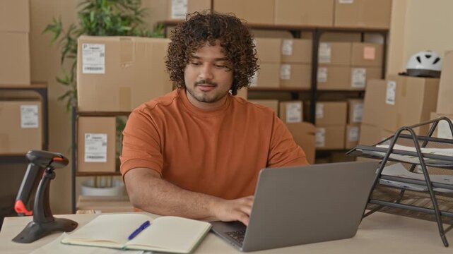 Man writing in green notebook beside laptop and stacked cardboard boxes inside a building shipping area; small business focused.