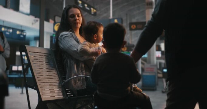 Young Family of Four Settles on Terminal Benches With Luggage. While Focused Father Checks Digital Boarding Passes and Travel Documents on Smartphone, Mother Attends to Two Small Children. Family