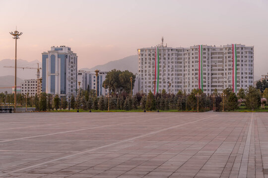 Independence Square in Dushanbe, Tajikistan, features vast paved plaza with cypress trees, modern high-rises with flag stripes, and Fann mountains backdrop.
