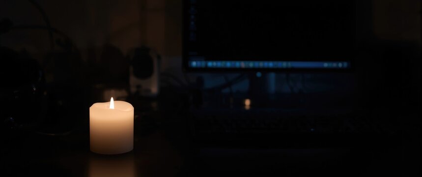 White candle creating a cozy glow on a desk with a dim and blurred computer in blackout