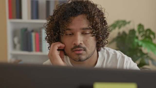 Man touching his cheek while looking intently at a computer monitor with a yellow sticky note, books on a shelf and a potted plant visible behind him at a desk in building; fatigue focus.
