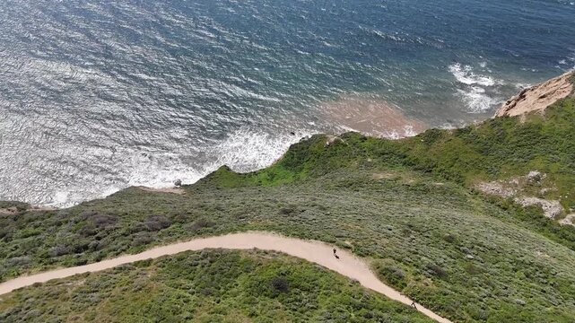 A stunning high-angle aerial shot captures a narrow dirt hiking trail winding along the edge of a steep coastal cliff. Two hikers are seen walking the path as the camera pans toward the turquoise ocea