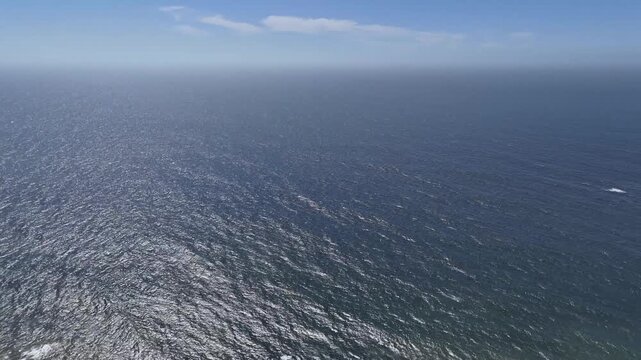 A sweeping aerial drone shot begins with the lush, pine-dotted hills of a coastal mountain range and pans smoothly to reveal the vast, shimmering blue expanse of the ocean. The footage captures the ru