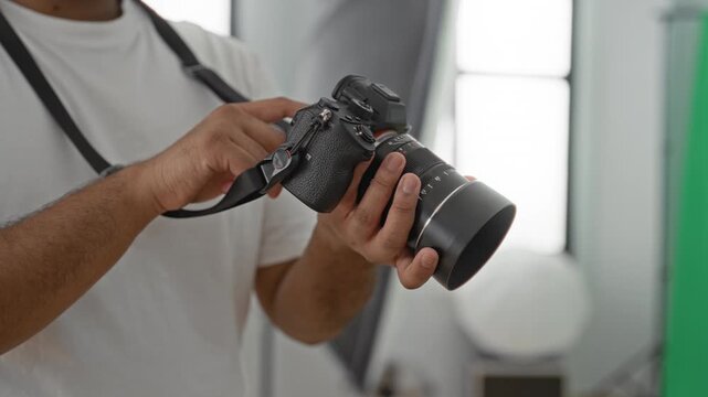 Young indian photographer adjusts camera dial with hands in studio near softbox and window; concentration precision craft.