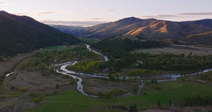Serpentine river channel weaving through green rural valley bordered by forested hills and distant mountains, aerial drone, ascend