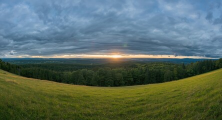 Obraz premium Sunlit meadow and forest panorama under a broad overcast blue sky