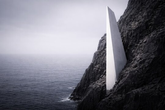 White minimalist geometric monolith on a dark rocky cliff overlooking a foggy ocean in a conceptual landscape