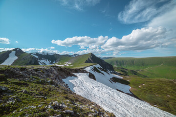 Scenic sunlit view along grassy ridge with white snow cornice to green hill top with rocky sheer crags in bright sun under lush clouds in blue sky. Vivid alpine scenery in high mountains in sunny day. © Daniil