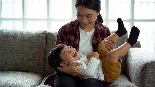 Happy Asian mother playing with her laughing baby son on the sofa at home family bonding and parenting concept joyful child playing on mother lap motherhood love care indoor