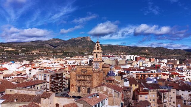 Aerial view of Chelva showing historic church tower, dense tiled rooftops and compact streets framed by Valencian mountains under scattered clouds