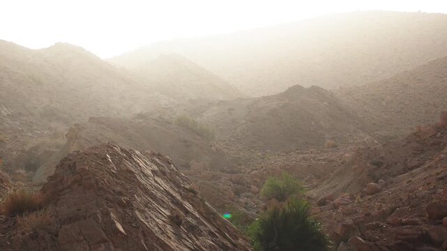 Aerial drone view of dramatic rocky mountain terrain with rugged layered rock formations and a narrow valley in Tunisia bathed in warm golden hazy light with sparse green vegetation