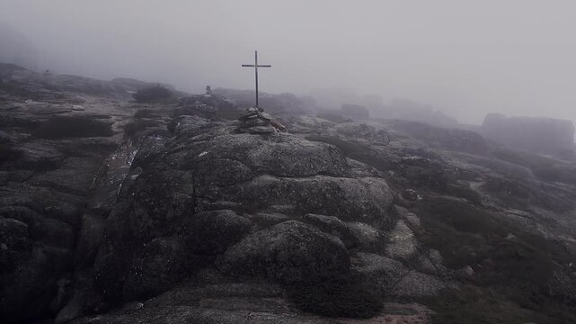 Summit cross stands on rugged granite ridge in Serra da Estrela Portugal surrounded by dense fog and harsh mountain terrain as drone pulls out and descends revealing wider landscape, aerial pull out