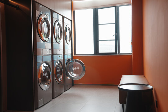 Modern self-service laundry room with multiple front-loading washing machines against orange walls near windows