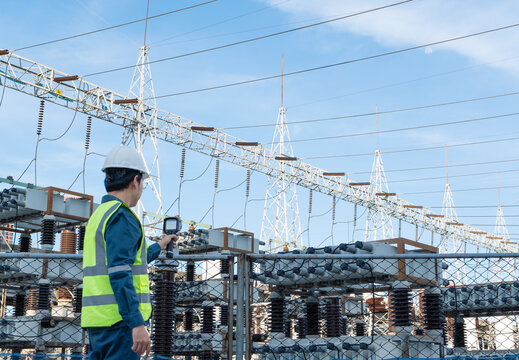 Engineer performs preventive maintenance with a thermal camera in a high voltage substation, checking outdoor equipment across a safety fence to support reliability and safety.
