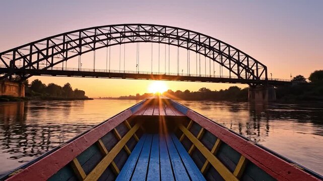 A long, narrow boat travels down a wide river towards a large arched bridge during a vibrant sunset