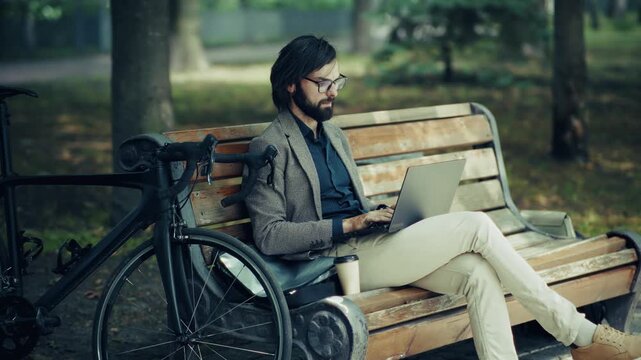 Freelancer Working On Laptop Sitting On Park Bench With Bicycle. Modern Technology