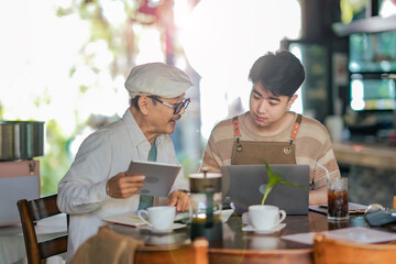 Fototapeta premium Asian father and Gen Z son analyze cafe startup data on laptop. SME family business focusing on accounting, financial planning and growth strategy in cozy coffee shop environment.