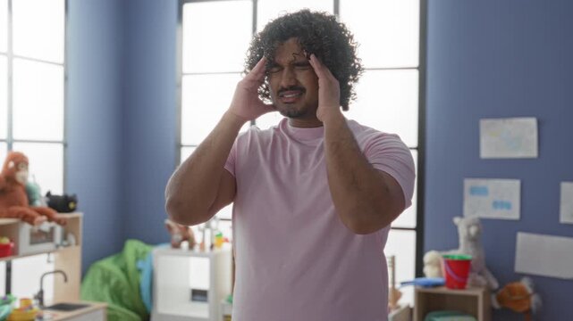 Young indian man rubbing forehead beside toys and a stuffed animal in a kindergarten building, holding temple in a tired pose; fatigue empathy.